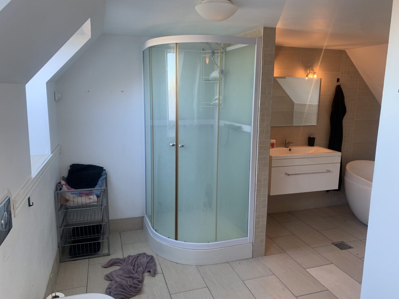 Investeringsejendom i Christiansfeld - Lyst badeværelse med hvide vægge og lyse fliser, der er en bruserkabine, håndvask og et spejl